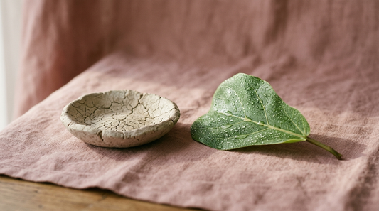 Dry cracked clay dish beside a dewy green leaf on dusty rose linen, illustrating dehydrated versus hydrated skin